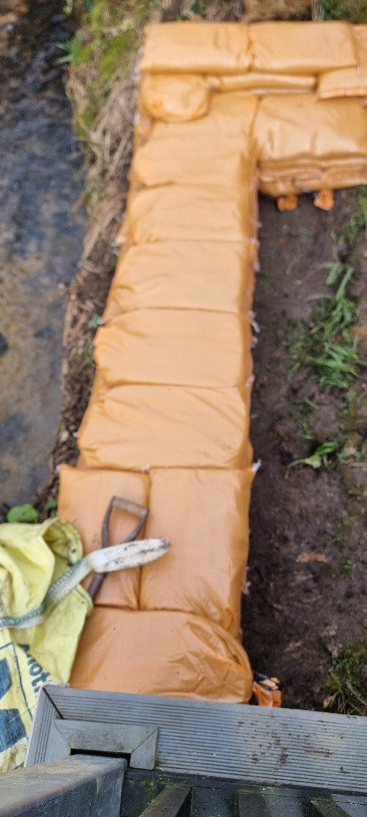 A brookside work area with tools and beige heavy-duty sandbags carefully positioned to create a stable flood prevention barrier.